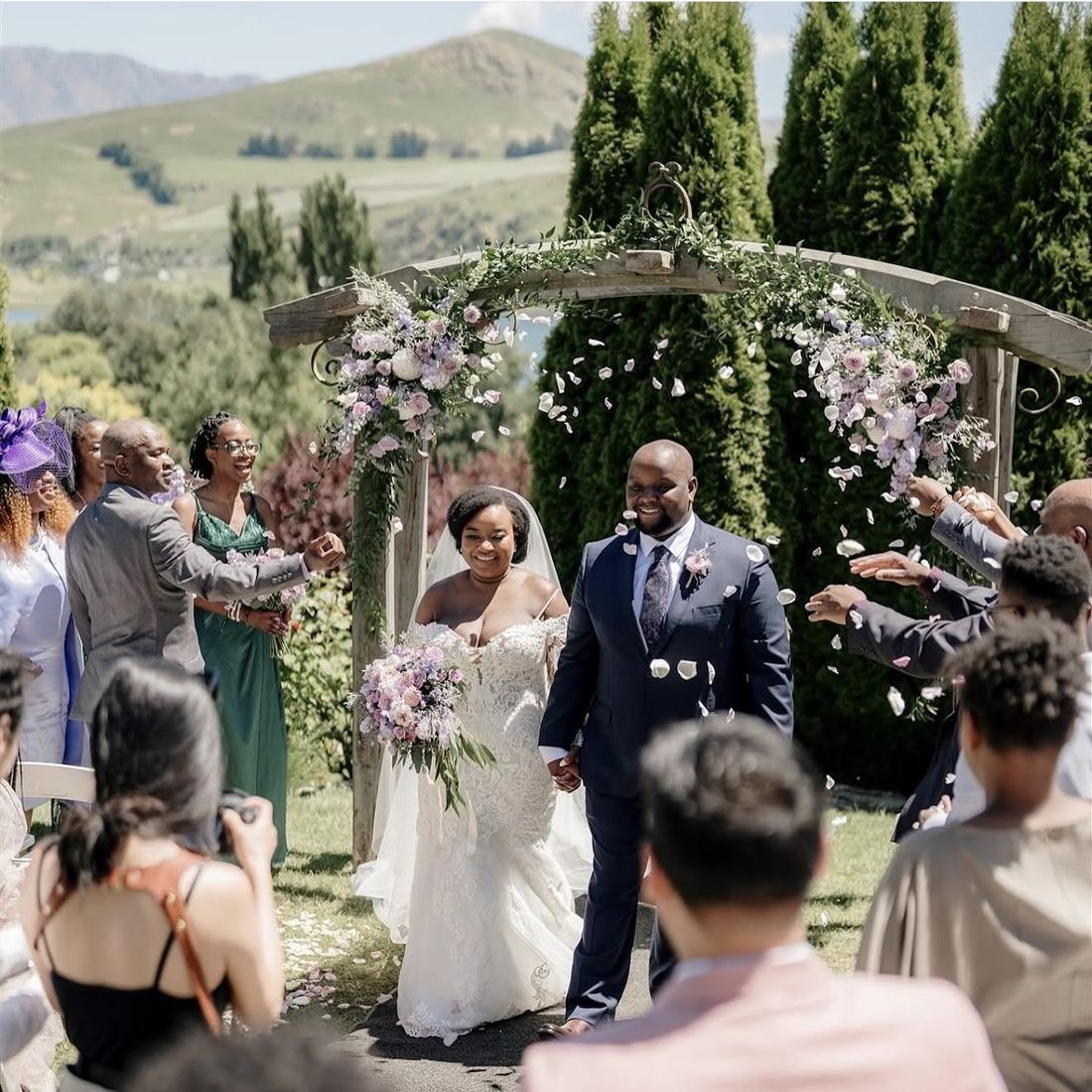 Bride and groom walking under a flower arch as guests throw petals