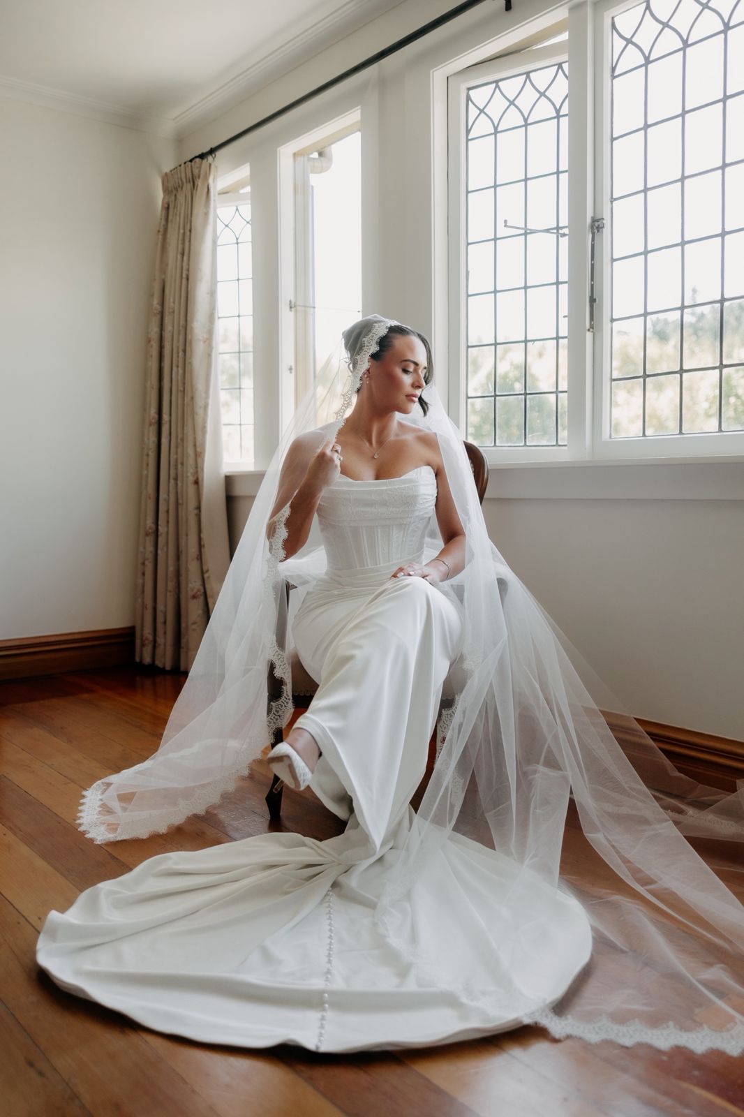 Bride seated by leadlight windows with a long lace-edged veil